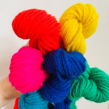 Load image into Gallery viewer, A hand holding a bundle of brightly colored yarn skeins in red, yellow, pink, blue, green, and teal against a plain light background.