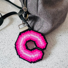 Load image into Gallery viewer, A close-up of a silver textured shoulder bag with a black strap, featuring a large, pink and white neon light style cross stitch letter 'C' bag charm with a black border, resting on a speckled light gray surface.