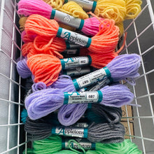 Load image into Gallery viewer, A white wire basket filled with multiple skeins of wool in assorted bright colors, including yellow, pink, orange, purple, gray, and green. Each skein has a label wrapped around it with the brand name Appletons visible.
