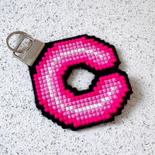 Load image into Gallery viewer, A pink and white neon light style cross stitched letter 'C' keychain with a black border and a silver metal key ring, placed on a speckled white surface.
