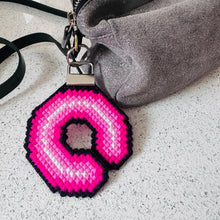 Load image into Gallery viewer, A close-up of a gray suede handbag with a black strap and a bright pink and white cross stitched letter 'C' bag charmcharm attached, resting on a light speckled countertop.

