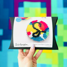 Load image into Gallery viewer, Hand holding a cross stitch coaster kit with colorful design against a multicolored geometric background
